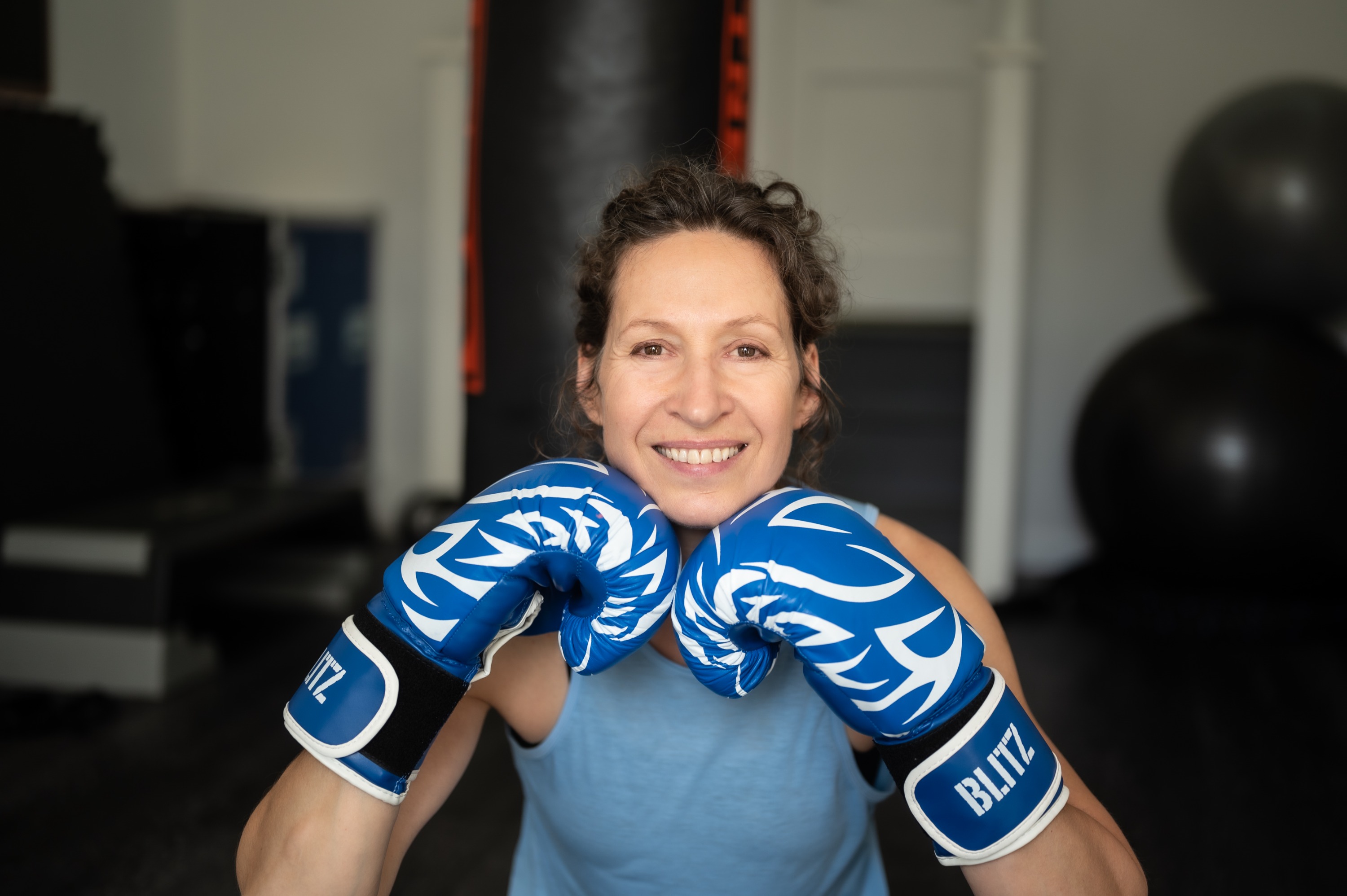 Tanya laughing, chin resting on two blue boxing gloves, in her private gym-studio