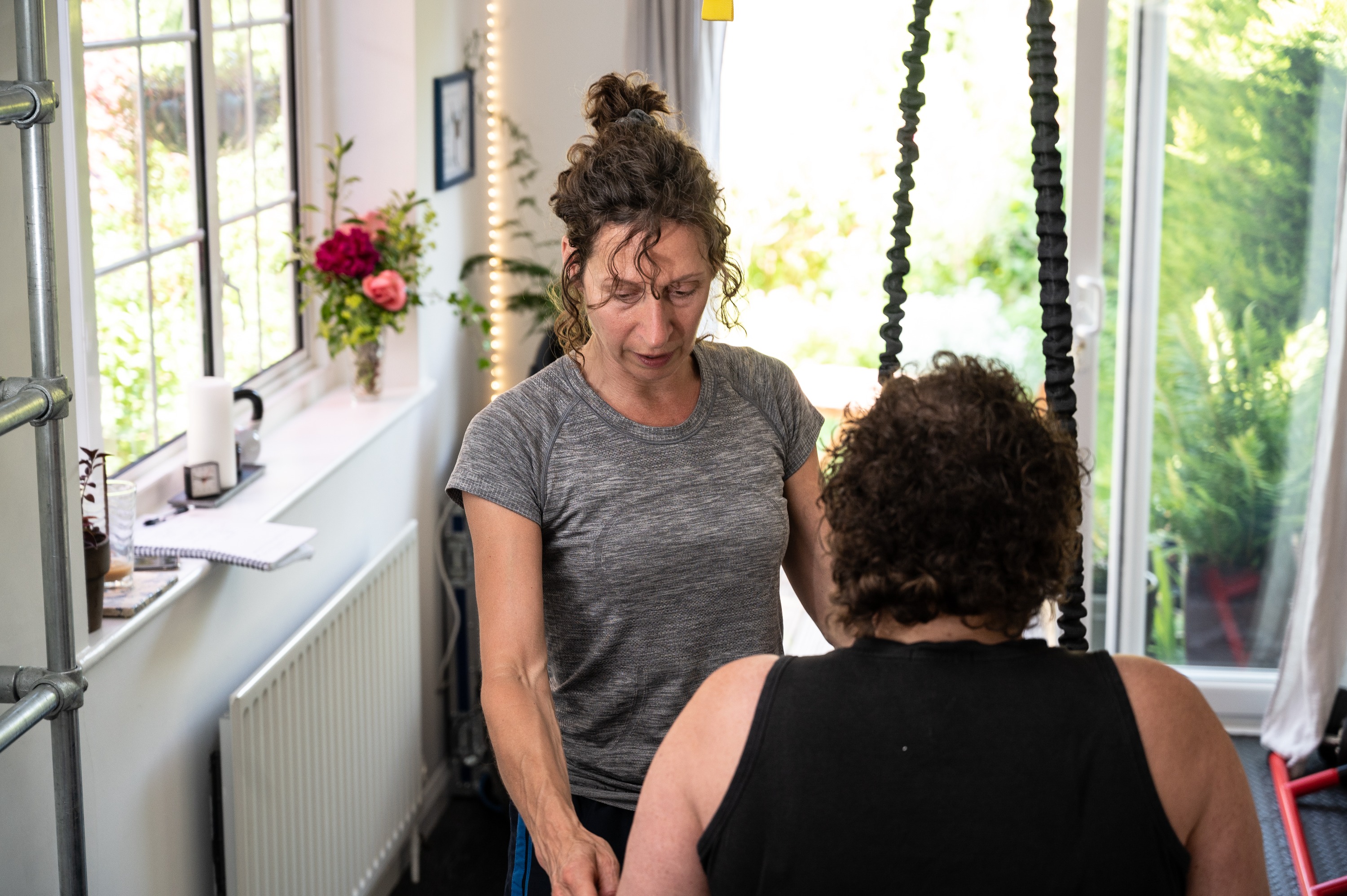 Tanya coaching a client beside TRX straps, garden doors and greenery behind, a jug of roses on the windowsill